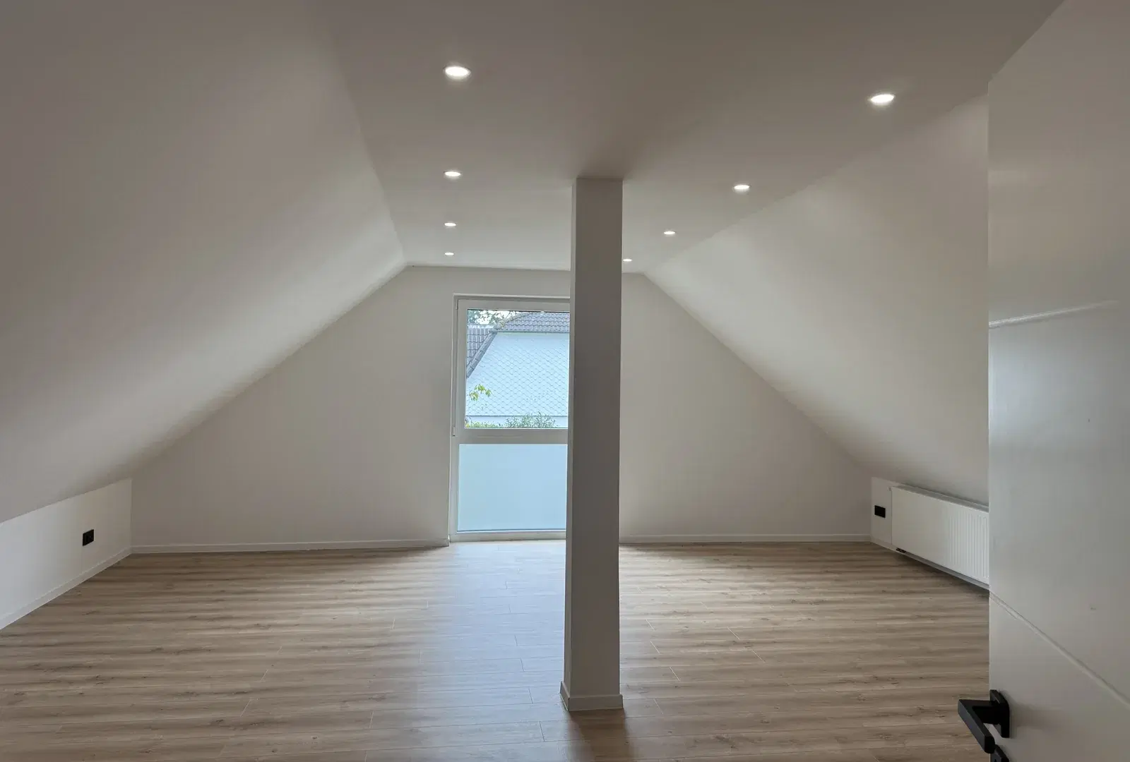 Fertig ausgebauter heller Wohnraum unter dem Dach in Hamburg-Poppenbüttel mit weißen Dachschrägen, Einbauspots, einer Stütze in der Raummitte, Fenstertür, Heizkörper und hellem Boden in Holzoptik